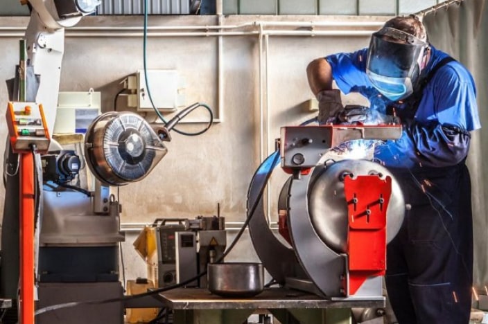 Man wearing a welding mask and polishing metal on a grinding wheel.